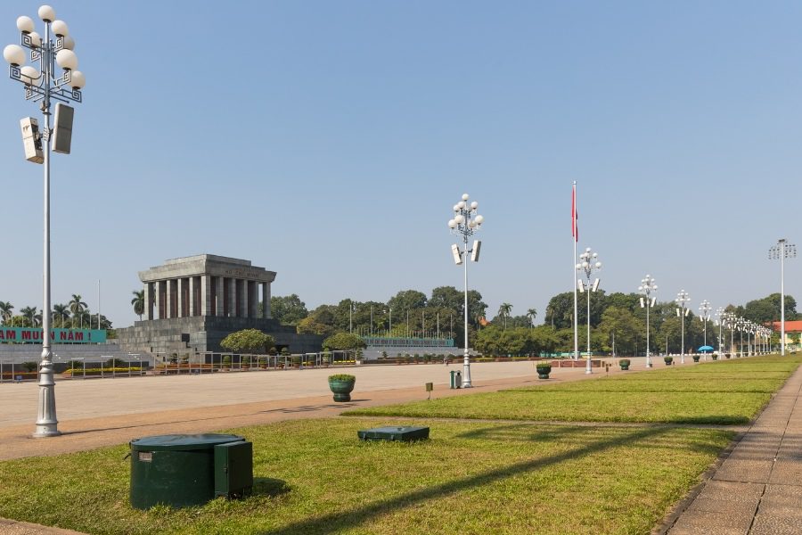 Wide view of a grand mausoleum in a large square with a Vietnamese flag, lined with ornate street lamps and lush greenery, a significant historical landmark.