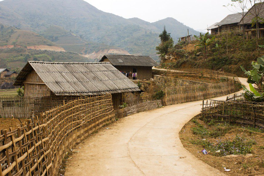 A rural village scene with traditional wooden houses topped with corrugated metal roofs, nestled along a winding dirt path. The area is surrounded by lush green hills and mountains, with bamboo fences lining the path and vegetation growing along the edges.