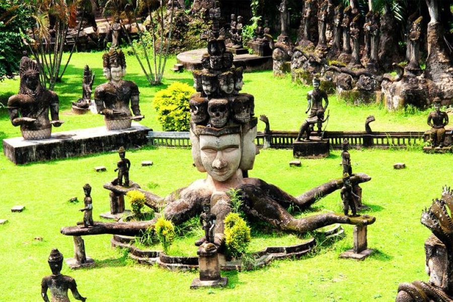 This is a photo of Buddha Park (Xieng Khuan) in Laos, showcasing a large, multi-faced deity statue surrounded by smaller mythological figures in a vibrant garden filled with Buddhist and Hindu sc