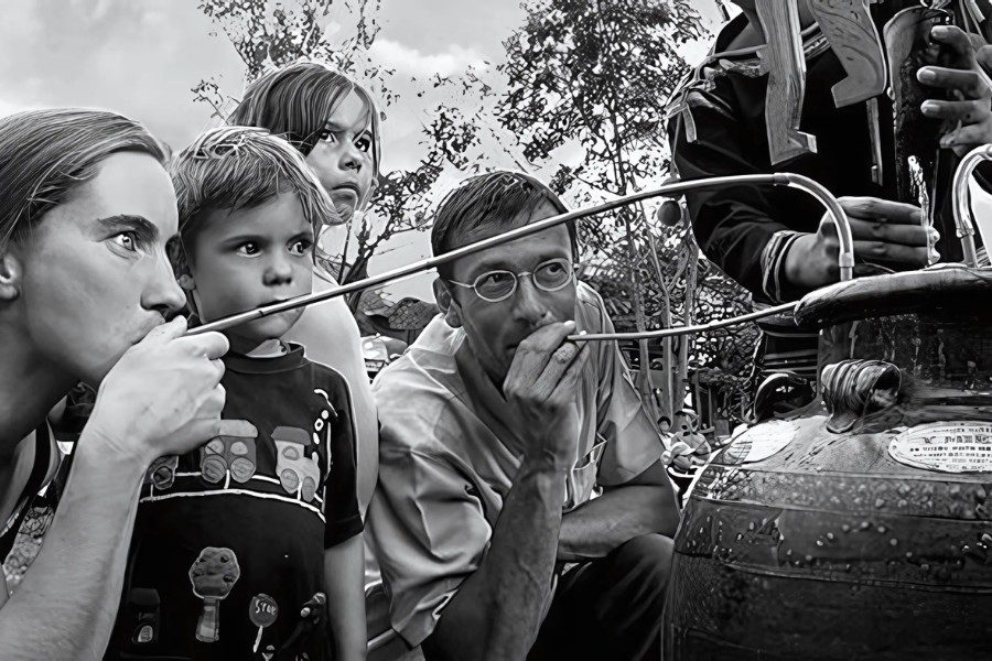 Children and adults curiously peering through tubes at a large metal container trying an uncommon drink, set against a natural backdrop.