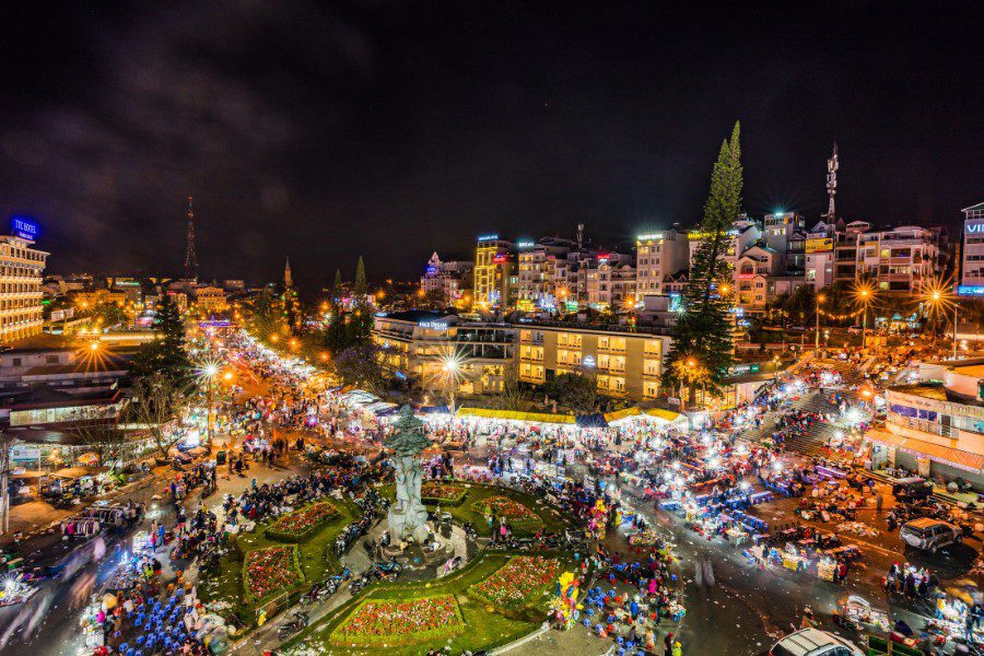 Vibrant night market in a bustling city square with colorful lights, a central statue, and numerous stalls, surrounded by illuminated buildings and a lively crowd, showcasing a dynamic urban scene.
