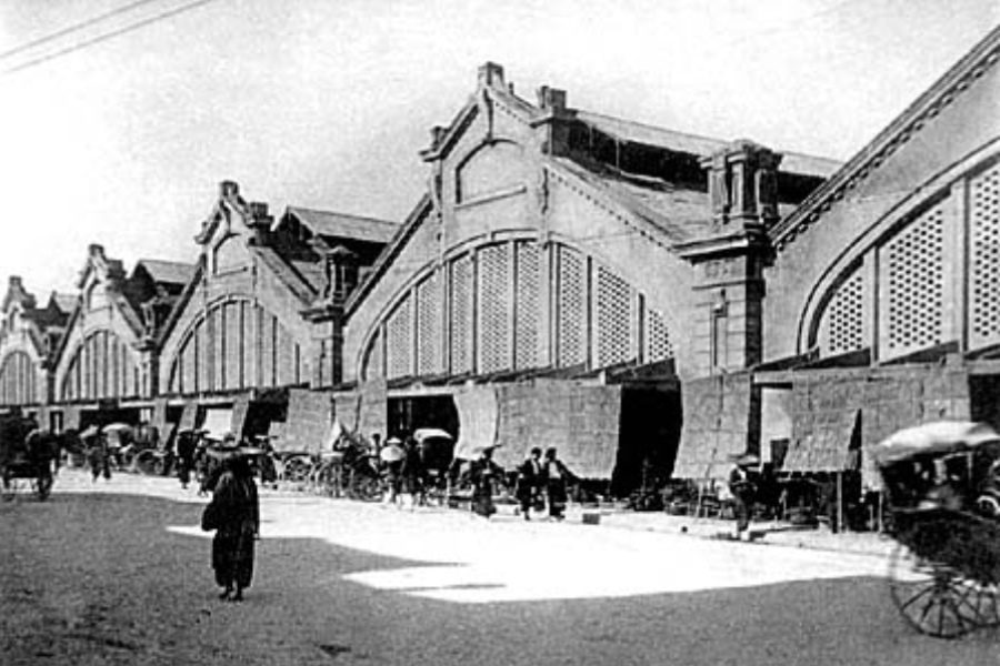 Black and white historical photo of a bustling market with arched architecture, featuring horse-drawn carriages and people in traditional attire, capturing a bygone era of trade and culture.