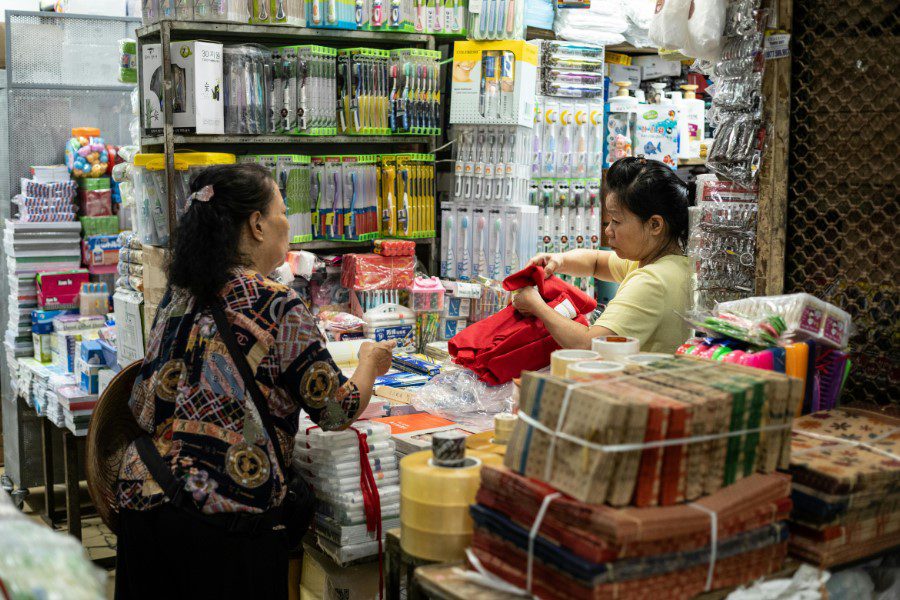 Two women shopping in a crowded market stall filled with various goods, including packaged items and household products, showcasing a vibrant local trading scene.