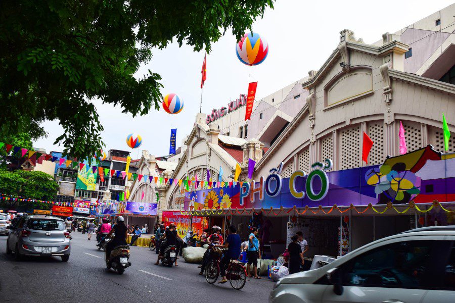 Vibrant street scene in front of a historic market adorned with colorful flags, balloons, and decorations, bustling with people, cars, and motorbikes, ideal for shopping and cultural tourism.