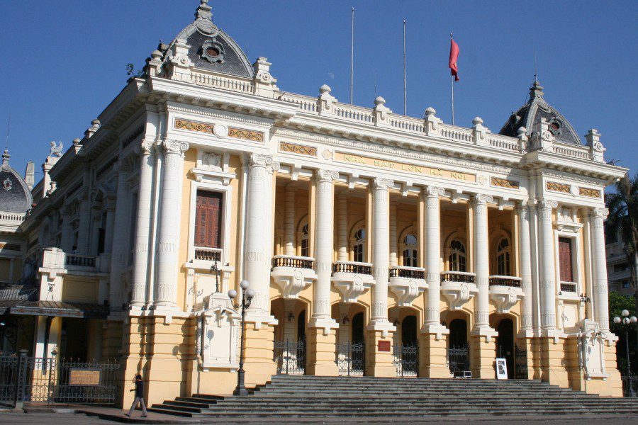 Grand colonial-style opera house with ornate columns and a Vietnamese flag, featuring intricate architecture under a clear blue sky, a cultural landmark in Vietnam.
