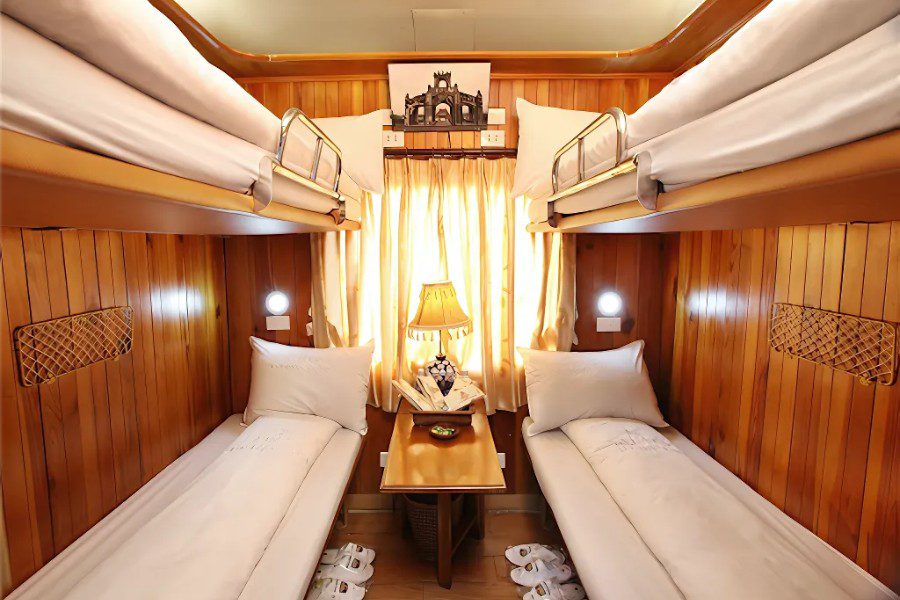 Cozy wooden cabin interior with two bunk beds, neatly made with white bedding, a small table with a lamp, and a window with curtains, offering a comfortable and inviting sleeping space.