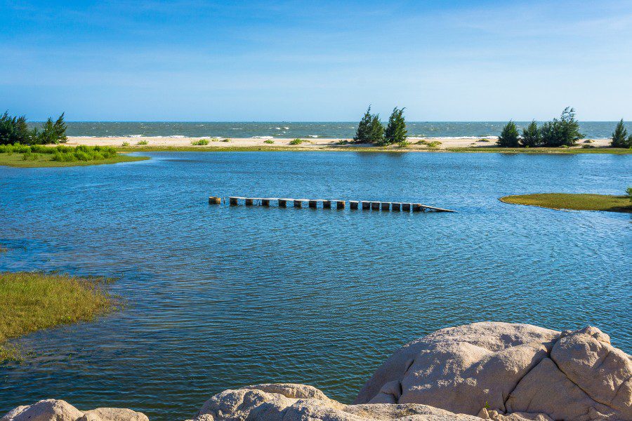 Scenic view of a calm lagoon with a small wooden bridge leading to a sandy shore, surrounded by lush greenery and pine trees, with the open sea and blue sky in the background, captured from a rocky vantage point.