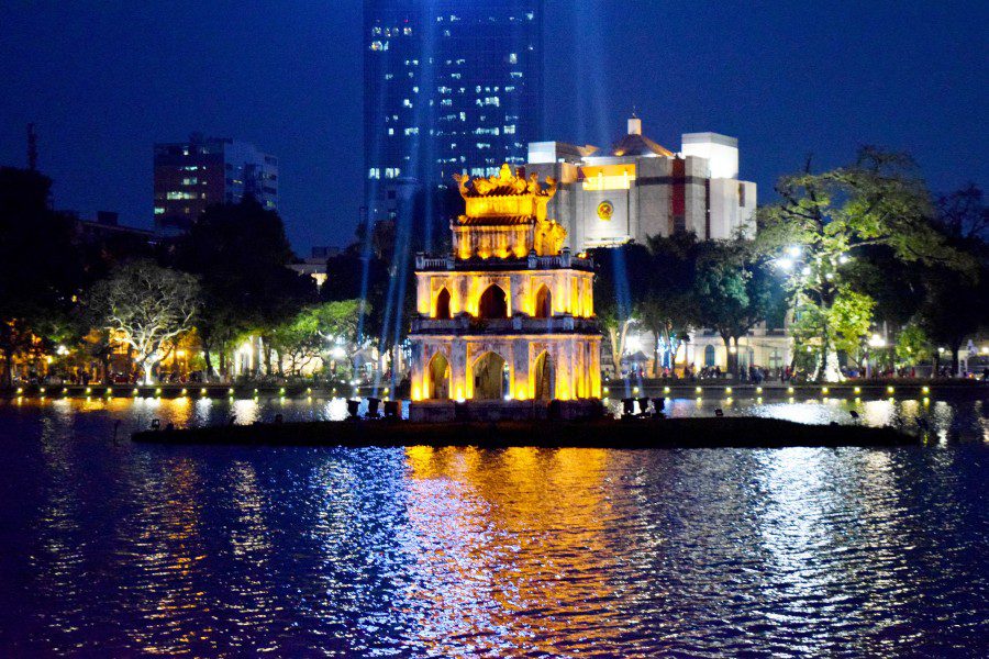 Iconic lit-up Turtle Tower on a small island in Hoan Kiem Lake at night, with reflections on the water and modern skyscrapers in the background, a must-see landmark in Hanoi.