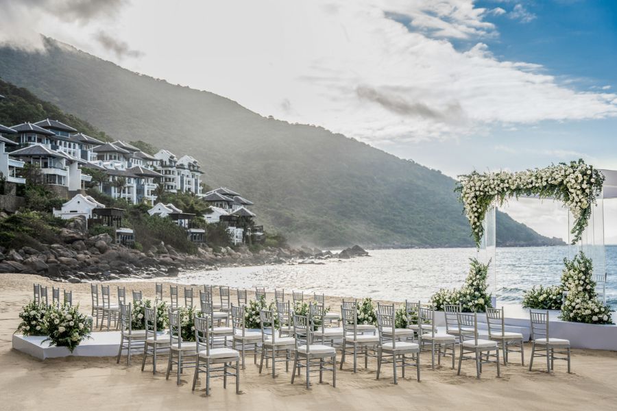 Beachfront wedding setup at a luxury resort in Vietnam, featuring elegant white chairs and floral arches, with ocean views and hillside villas in the background.