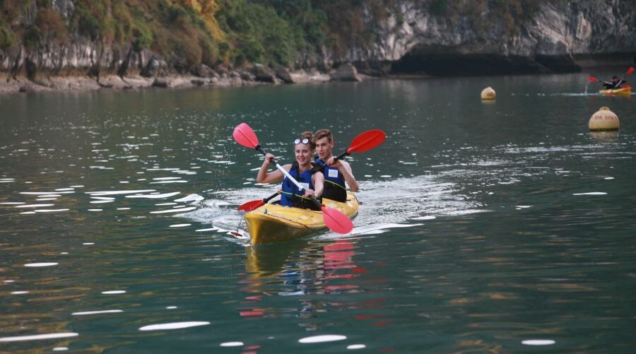 Two people paddle a yellow kayak in Halong Bay or Lan Ha Bay, surrounded by rocky cliffs.