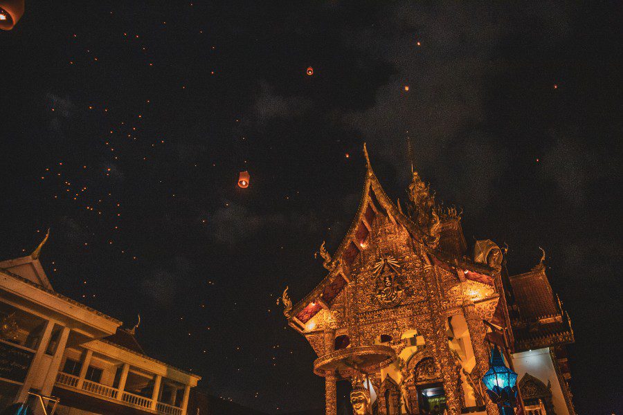 Loy Krathong night with a beautifully ornate Thai temple illuminated against a dark sky, glowing lanterns floating upwards, creating a magical atmosphere