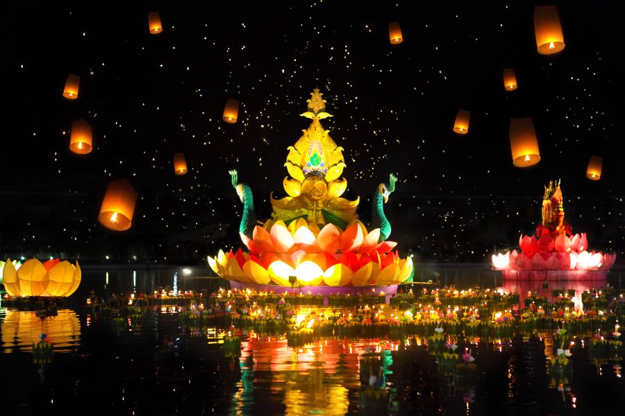 Glowing krathongs float on a river during the Loy Krathong festival Thailand under a full moon, with sky lanterns illuminating the night sky.