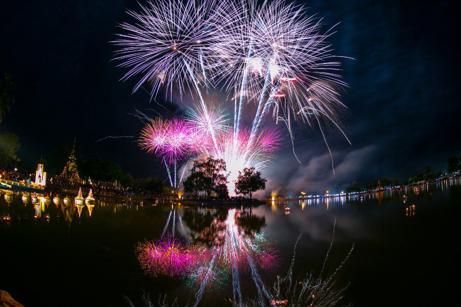 In Sukhothai in Thailand, on Loy Krathong day there will be fireworks display over a serene lake, reflecting vibrant purple, pink, and white bursts in the water, with a traditional Thai temple silhouetted on the shore