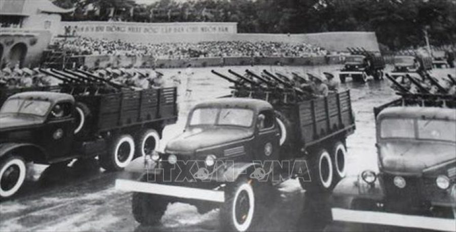 Black and white historical photo of military trucks carrying anti-aircraft guns, with a large crowd in the background, symbolizing a significant military parade.