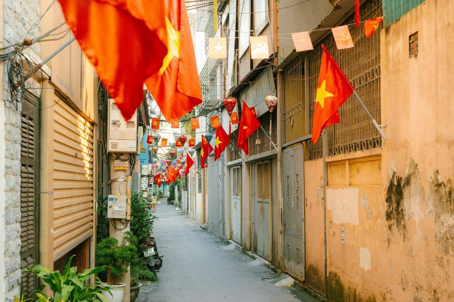 Narrow alleyway decorated with Vietnamese flags and lanterns, lined with traditional buildings and lush plants, capturing the vibrant cultural atmosphere of a national celebration.