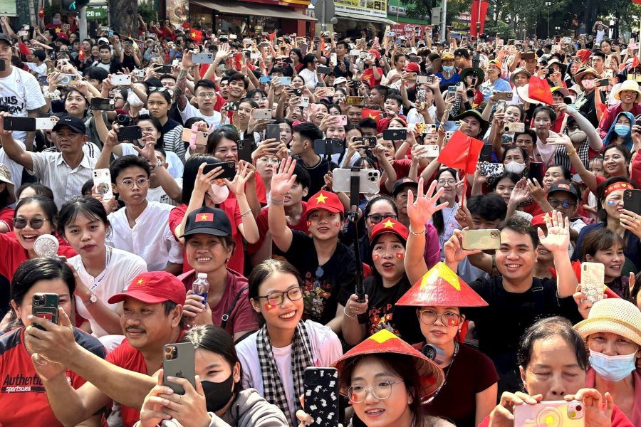 Large crowd celebrating with Vietnamese flags, taking photos, and waving, many wearing traditional hats and red attire, capturing a lively national event in Vietnam.