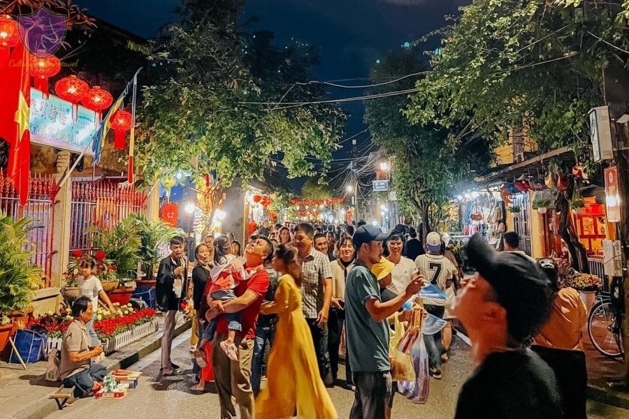A lively Vietnamese night market bustles with families, lanterns, and joyful energy under a canopy of trees and lights.