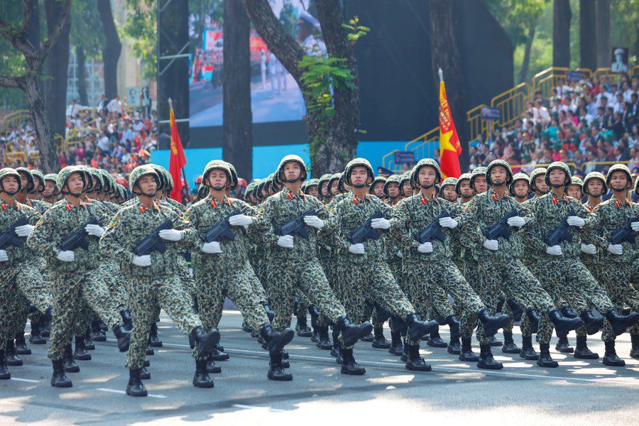 Military parade with soldiers in camouflage uniforms marching in unison, holding rifles, with Vietnamese flags and a large crowd in the background, symbolizing national pride and unity.