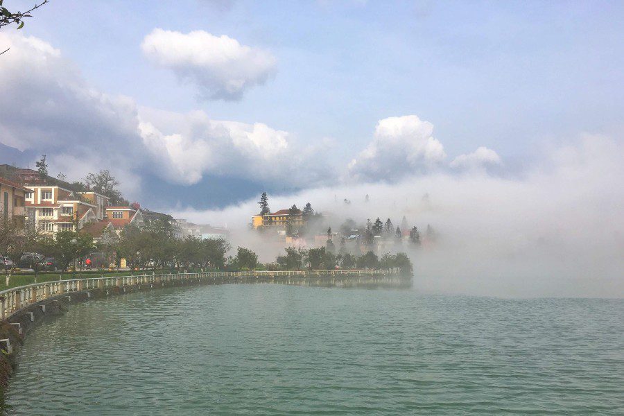 A peaceful lake scene with clear blue water, next to a white fenced walking path running along the shore. In the distance, houses and trees are shrouded in thick fog.