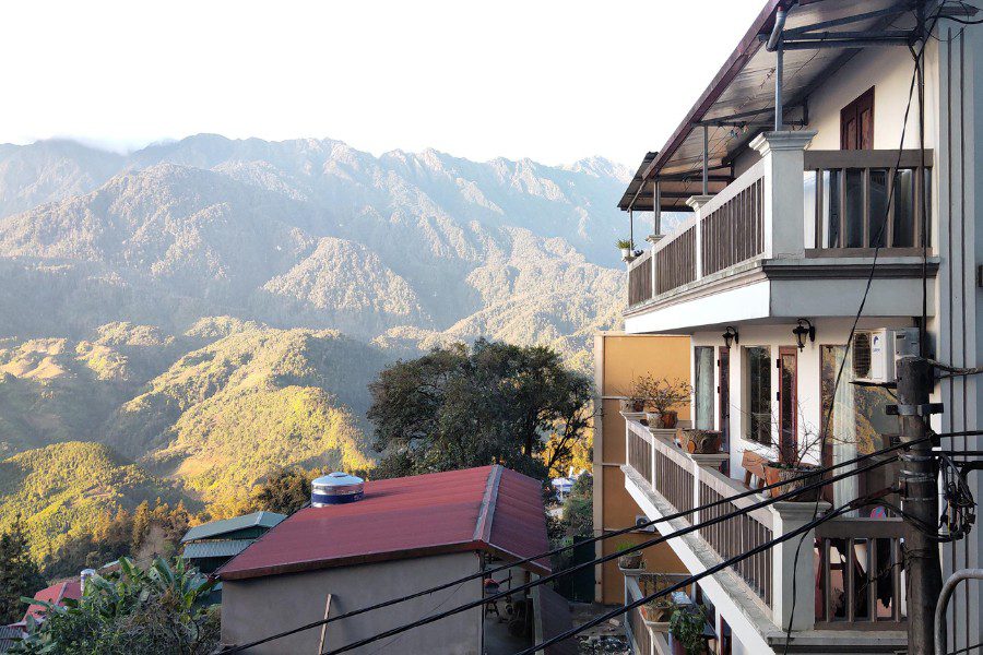 A rural scene with a two-story building with balconies and windows, next to a red-tiled roof. Surrounded by lush green mountains and valleys, with dense trees and some electric wires running through. The early morning light illuminates the landscape, creating a peaceful atmosphere.