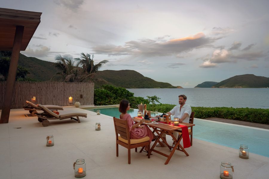 Romantic dinner setup at a beach resort in Vietnam, a couple dining by a private pool with candlelight, overlooking the ocean and distant mountains at dusk.