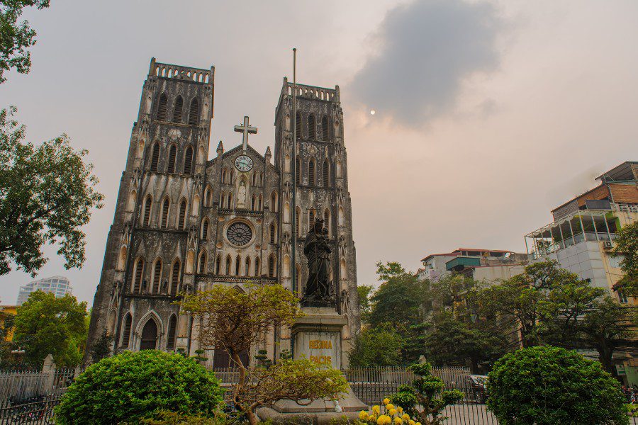 Historic St. Joseph's Cathedral with twin towers and a statue in front, surrounded by lush greenery and urban buildings under a cloudy sky, a prominent landmark in Hanoi.