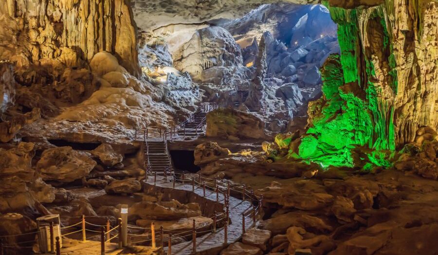 A large cave interior with stalactites and stalagmites, illuminated by artificial lighting. Wooden walkways and stairs with railings guide visitors through the cave, surrounded by rugged limestone structures and rocky terrain.