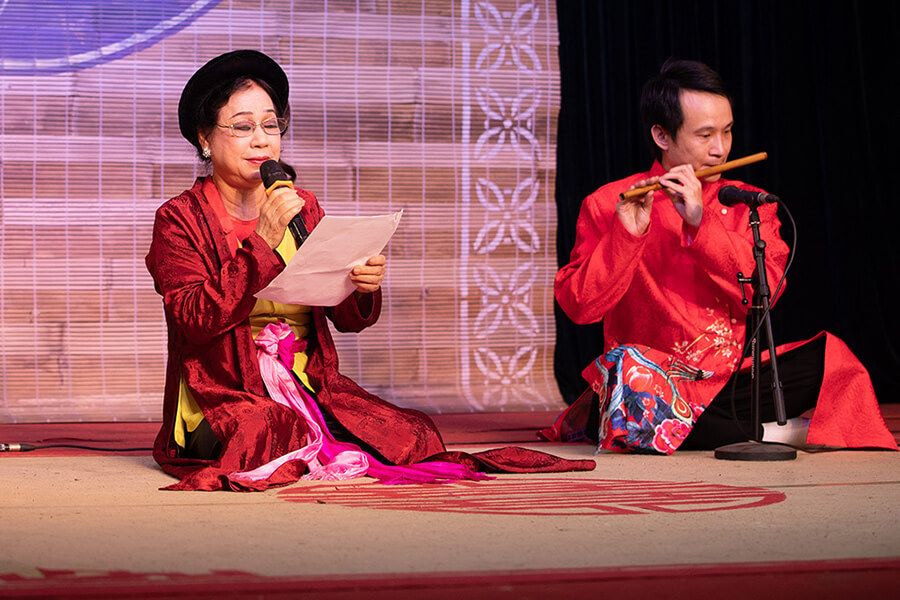 A woman sings while a man plays the flute in a traditional Vietnamese music performance, both dressed in vibrant áo dài, celebrating cultural heritage.