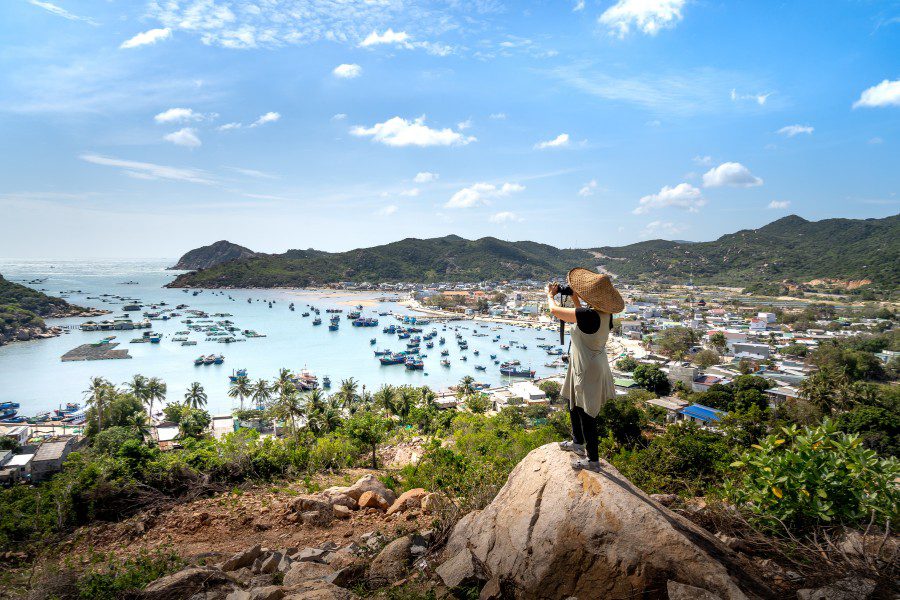 A person wearing a traditional conical hat stands on a rocky outcrop, taking a photo of a scenic coastal village with numerous fishing boats in a bay, surrounded by lush green hills under a clear blue sky.